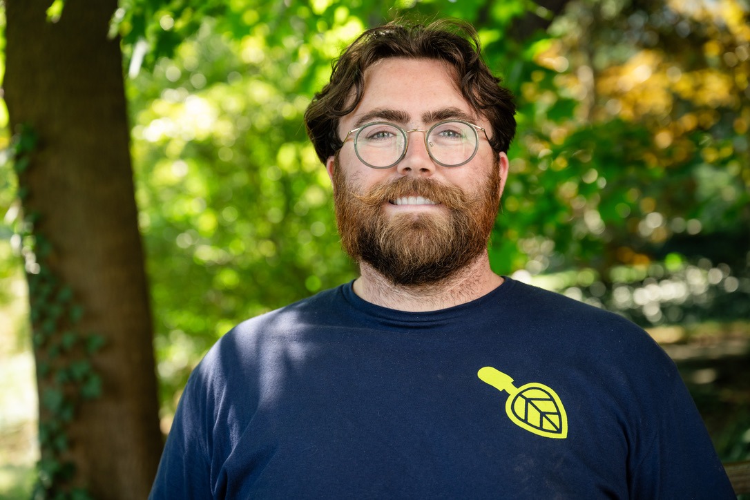 A person with glasses and a beard stands outdoors in a lush green setting, wearing a navy shirt with a yellow design.