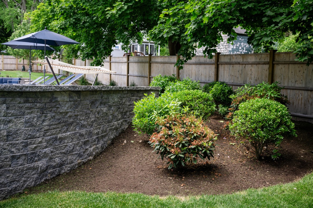 A garden area with neatly trimmed bushes, a stone wall, and a hammock near a wooden fence beneath large trees.