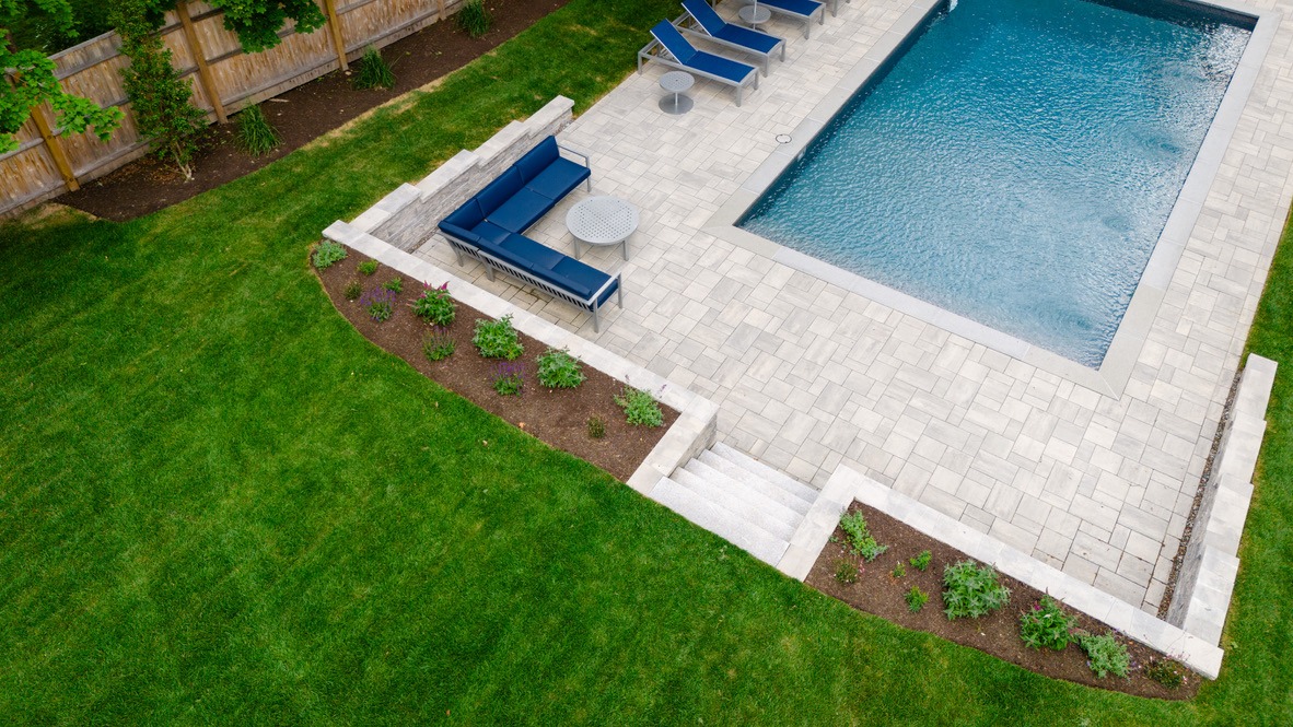 Aerial view of a backyard with a rectangular pool, surrounding patio, blue lounge chairs, L-shaped sofa, and neatly manicured lawn.