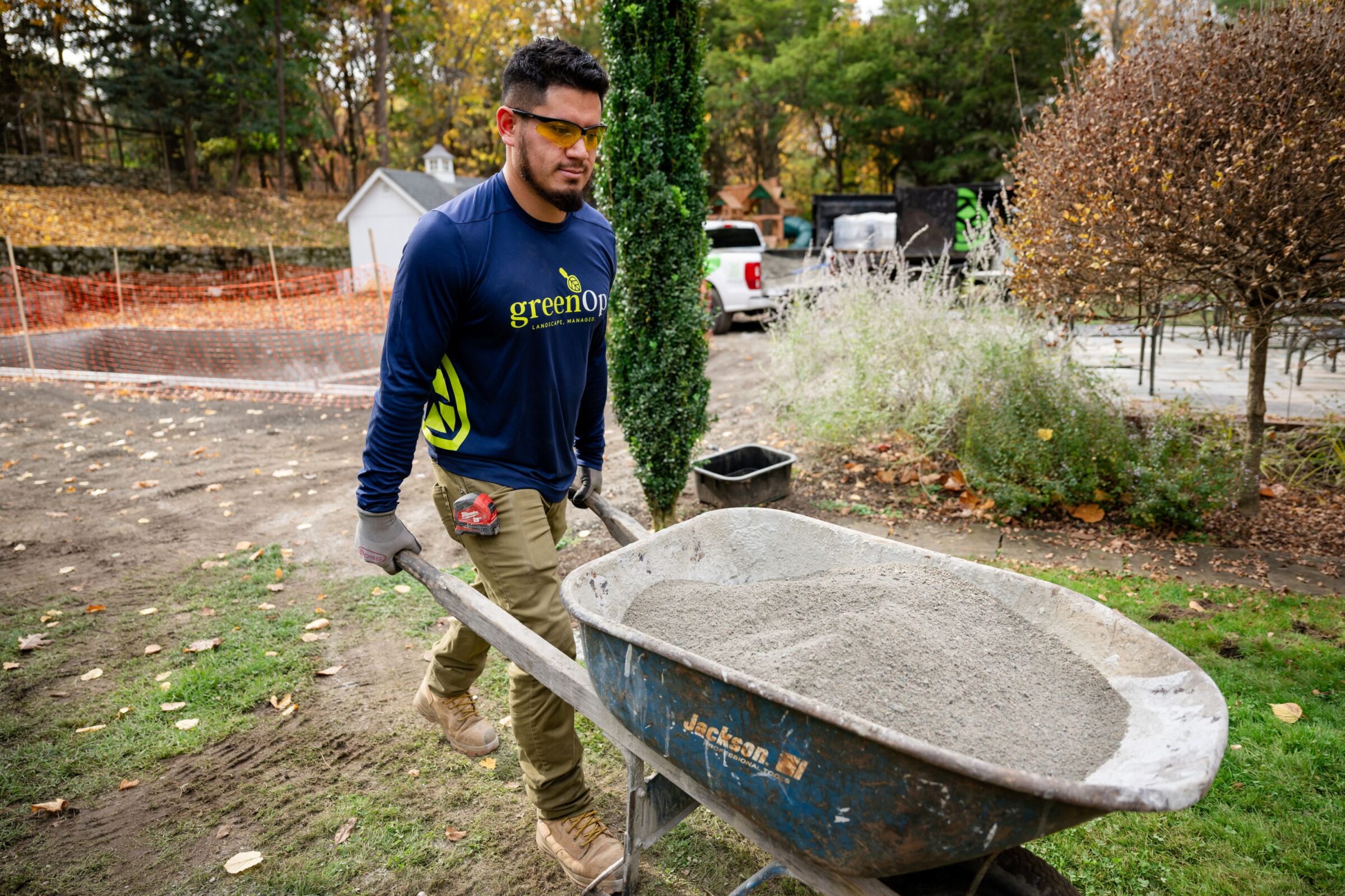 A person with protective eyewear moves a wheelbarrow of cement in a garden, surrounded by autumn foliage and construction materials.