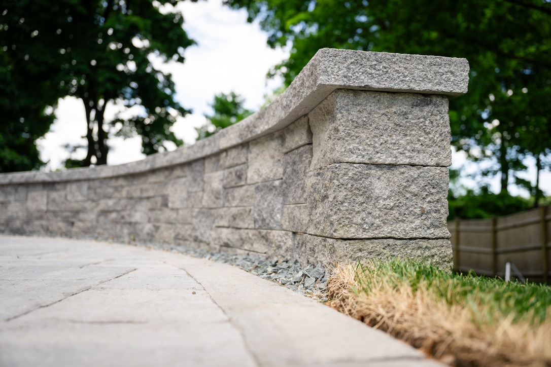 A stone wall curves through a park setting, surrounded by trees and grass, under a partly cloudy sky.