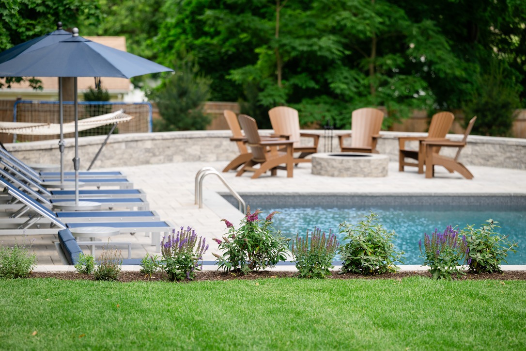 A serene backyard features a pool, lounge chairs, umbrella, vibrant garden, hammock, and firepit surrounded by wooden chairs, set against lush greenery.