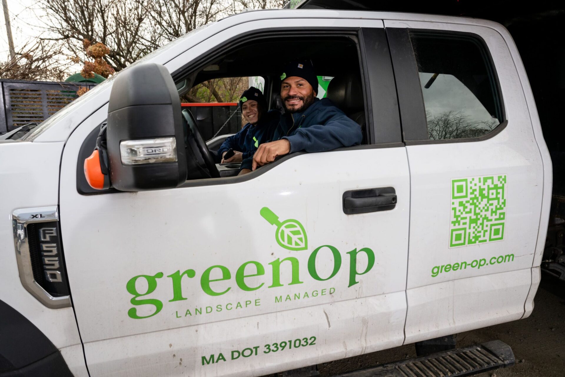 Two people smiling inside a white truck with "greenOp" logo, parked outdoors. The truck door displays a QR code and company information.