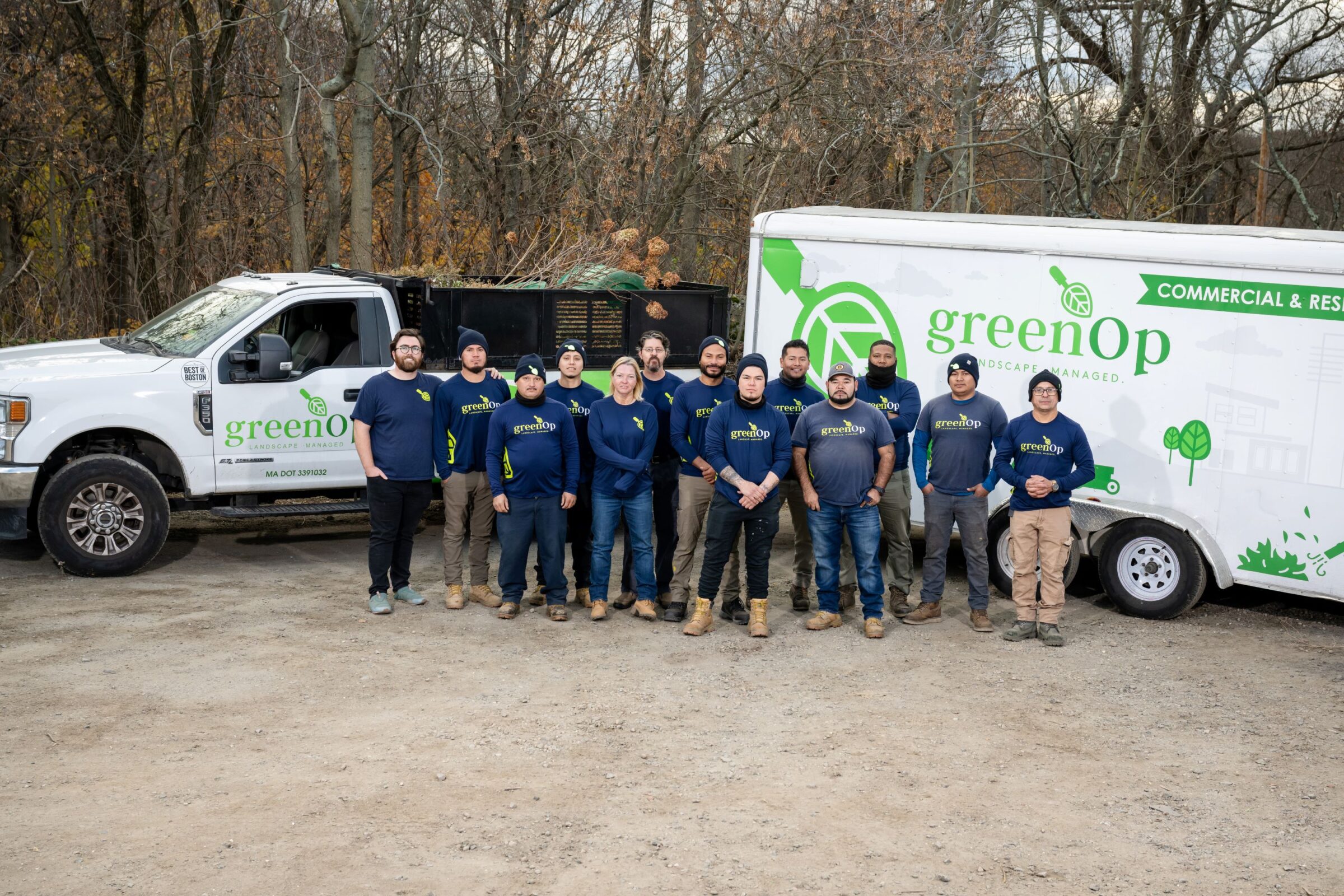 A group of people stands before a "greenOp" truck and trailer, wearing matching gear, amidst a wooded area.