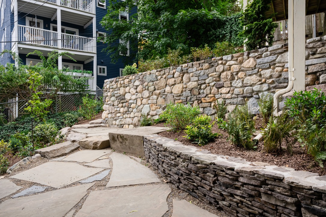 Stone pathway and retaining wall bordered by plants, leading to a multi-story blue building with balconies surrounded by greenery.