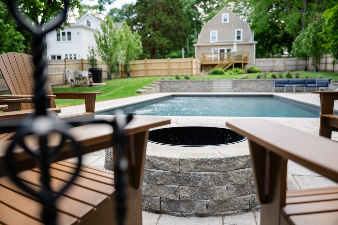 Backyard scene with wooden chairs, a round stone fire pit, and rectangular pool. House and trees visible in the background.