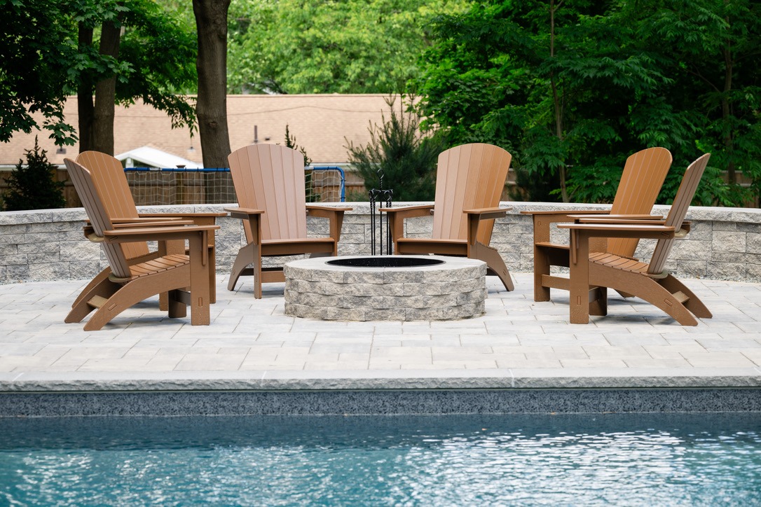 Four Adirondack chairs surround a stone fire pit on a patio by a pool, with trees and house in the background.