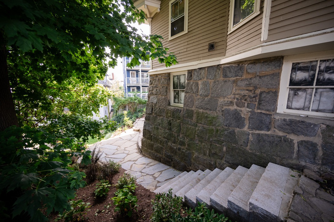 Stone steps lead down beside a house with a stone foundation. Greenery surrounds the path, and additional buildings are visible.