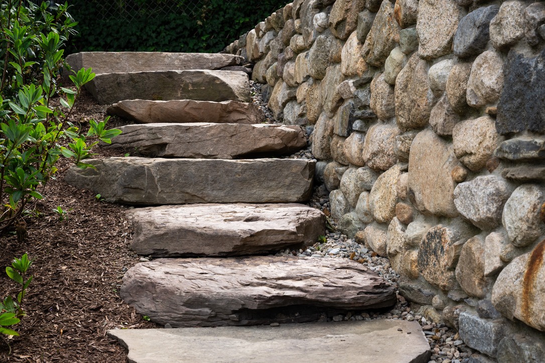 Stone steps ascending beside a natural stone wall, surrounded by lush greenery, creating a peaceful garden atmosphere.