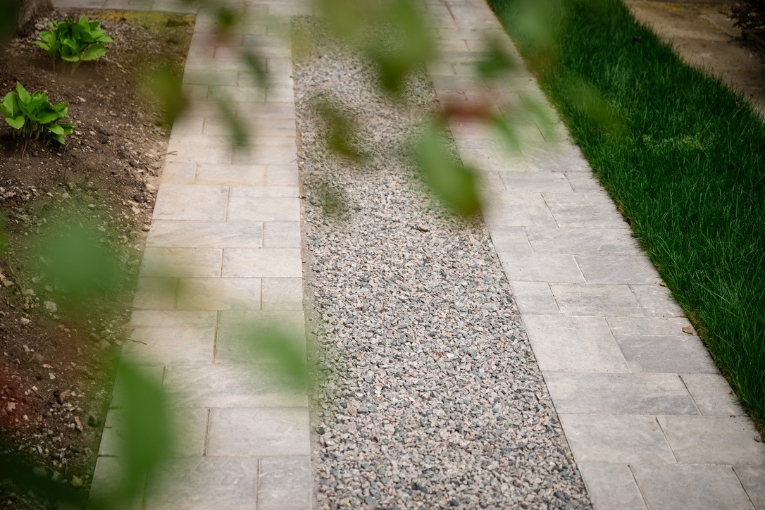 A stone and gravel path lined with green grass and plants, partially obscured by blurred foliage in the foreground.