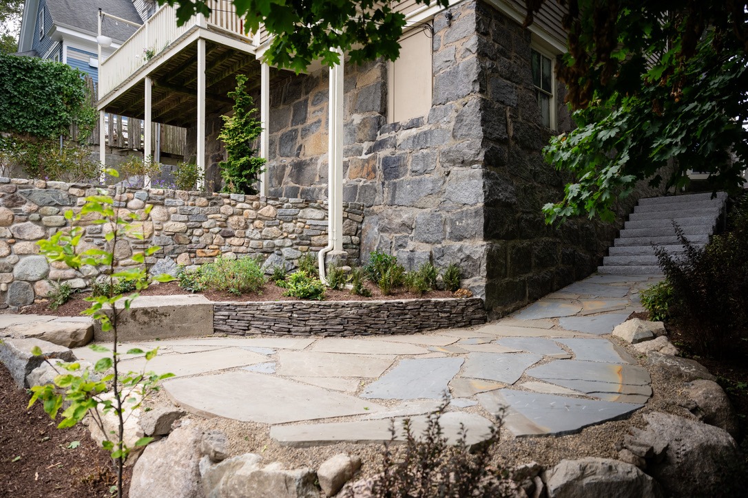 Stone house with textured walls, surrounded by landscaped garden and flagstone path, leads to a set of outdoor stairs. Lush greenery accents scene.