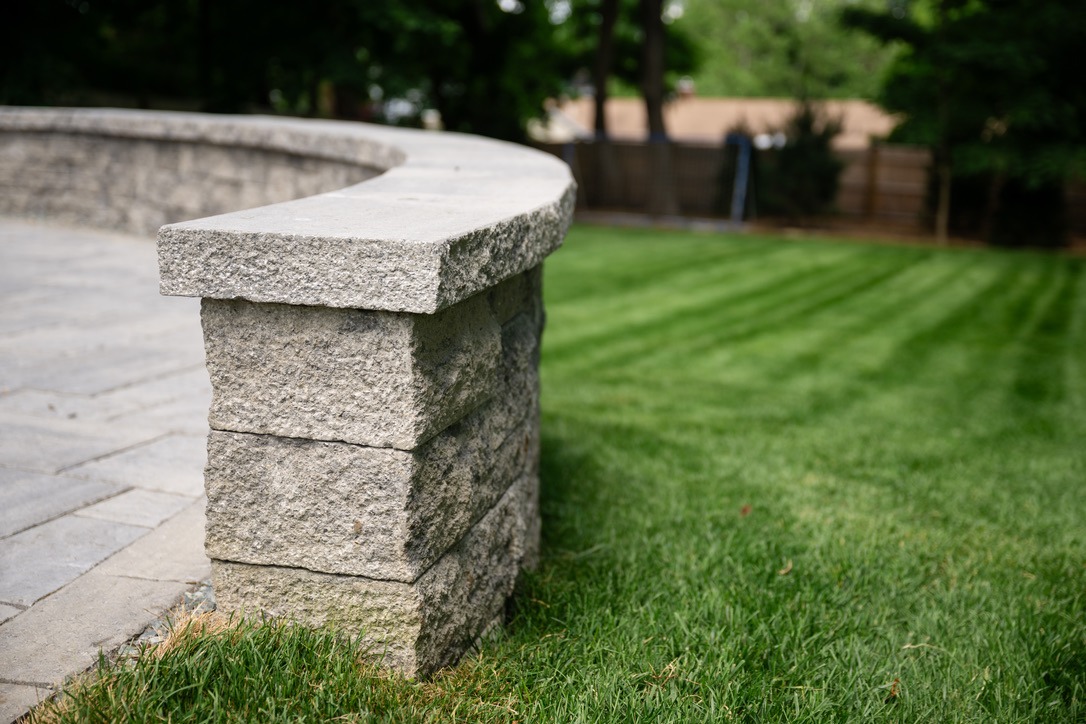 Curved stone wall on a well-manicured lawn, bordered by greenery and trees. No recognizable landmarks or historical buildings visible.