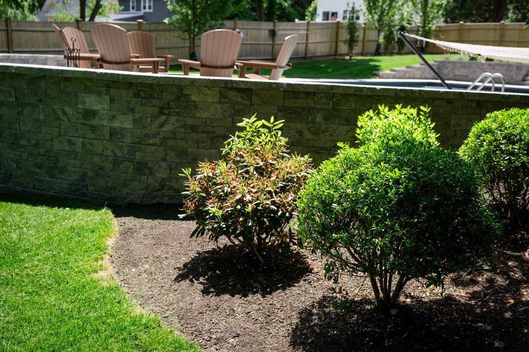 A backyard features a neatly trimmed garden, stone wall, Adirondack chairs, and a hammock, with trees and a wooden fence in the background.