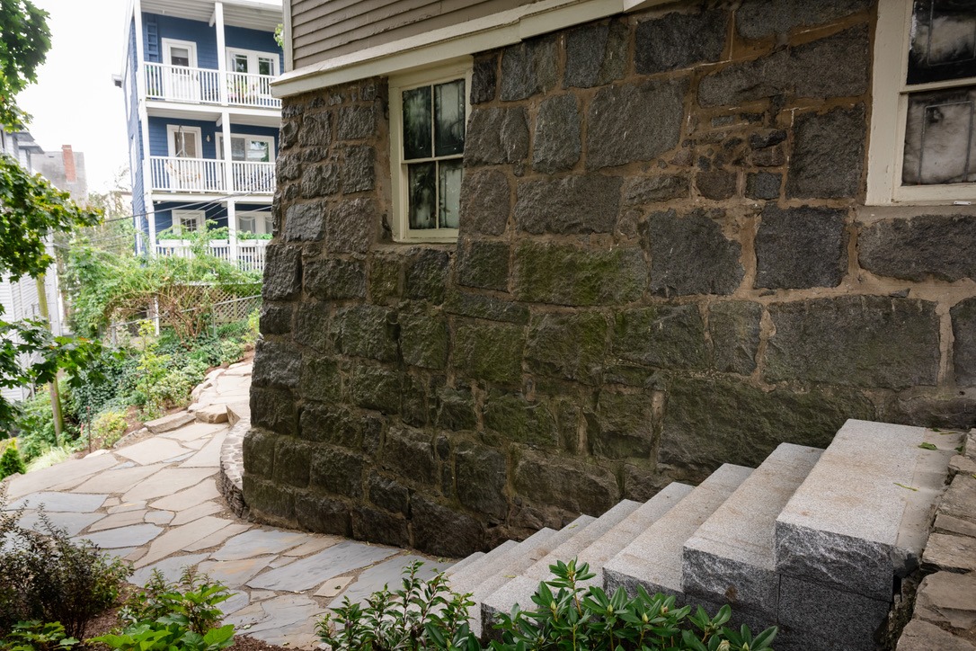 Stone steps lead to a historic stone building with vintage windows, surrounded by greenery. A wooden house with balconies in background.