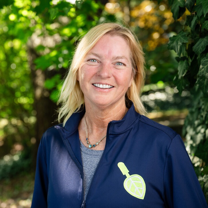 A person with blonde hair smiles outdoors, wearing a navy jacket with a leaf logo. Green foliage is visible in the background.