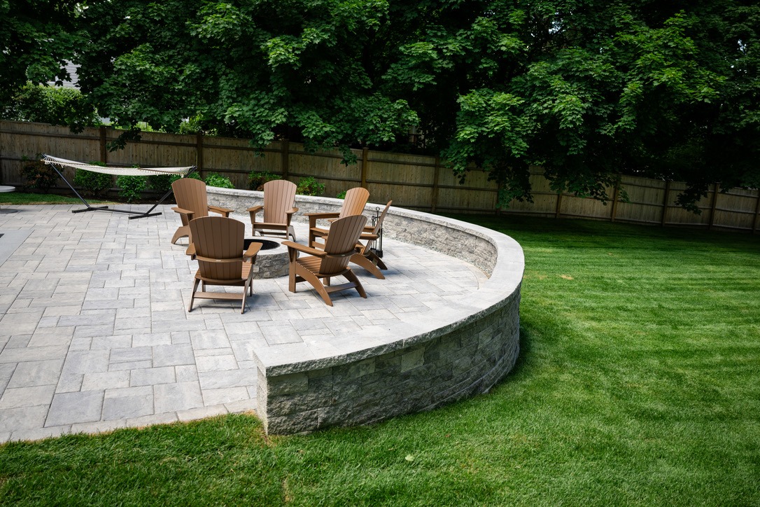 A backyard patio with five wooden chairs, stone wall, hammock, and green grass, surrounded by trees and a wooden fence.