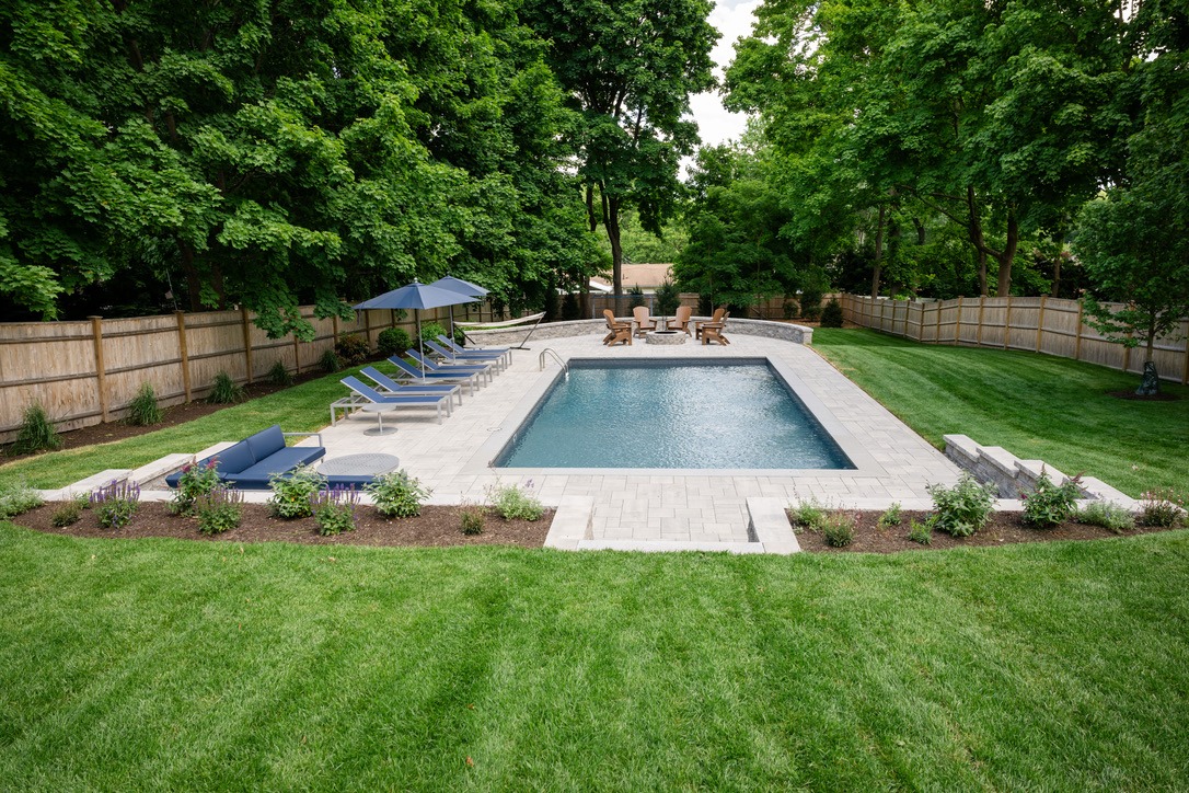 Rectangular backyard pool with lounge chairs, umbrellas, surrounded by landscaped greenery and trees, bordered by a wooden fence and patio seating area.