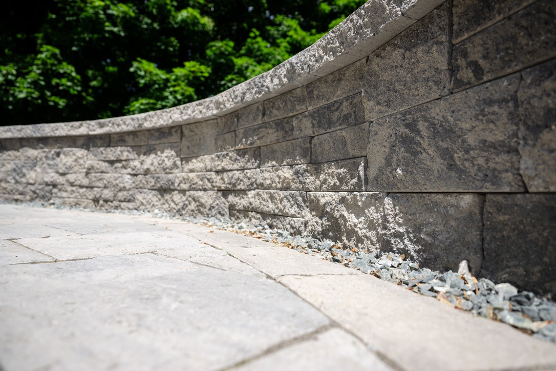 Curved stone wall alongside paved path with small rocks, flanked by lush green trees in the background under bright sunlight.