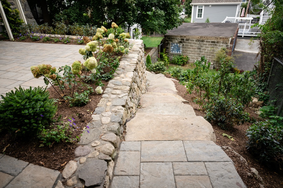 Stone path with adjacent flower beds and shrubs, leading to a garden area near a brick building and a detached garage.