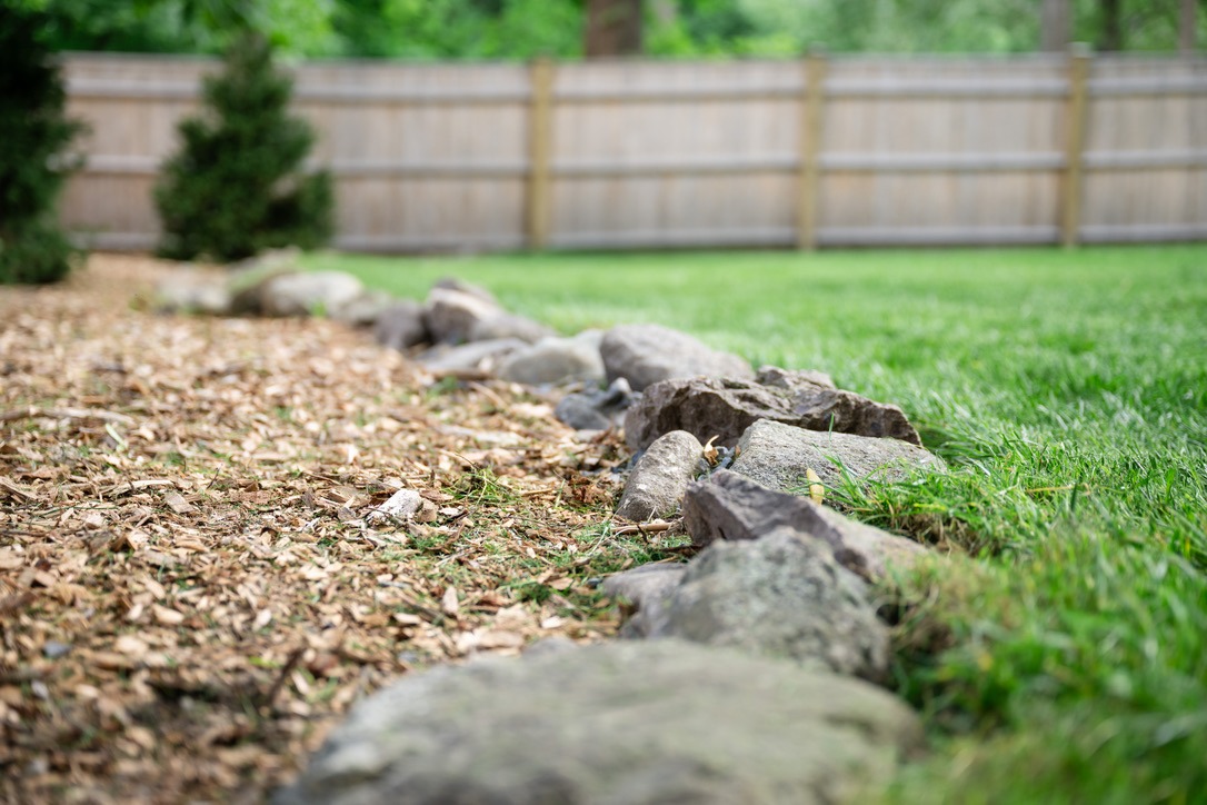 A garden path of wood chips is bordered by rocks, with grass on the side and a wooden fence in the background.