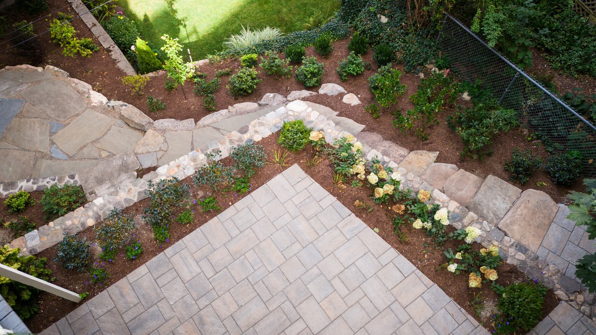 Aerial view of a landscaped garden with paved patio, stone pathway, and neatly arranged plants, surrounded by greenery and fencing.