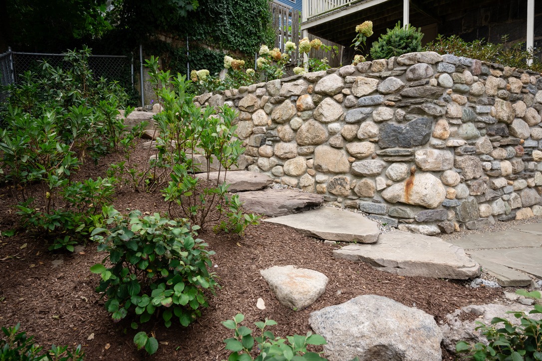 Stone pathway with lush green plants leads to a raised garden surrounded by a rustic, stacked stone wall under a weathered wooden structure.