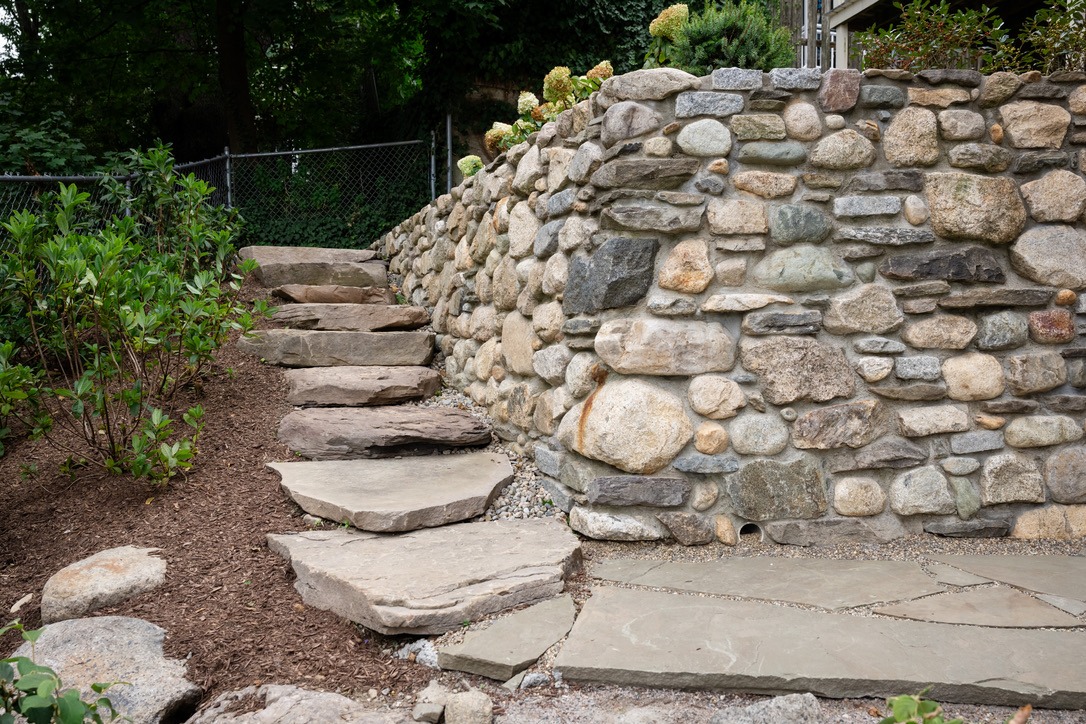 Stone pathway leads up stone steps beside a rustic stone retaining wall and garden with green foliage, against a backdrop of trees.