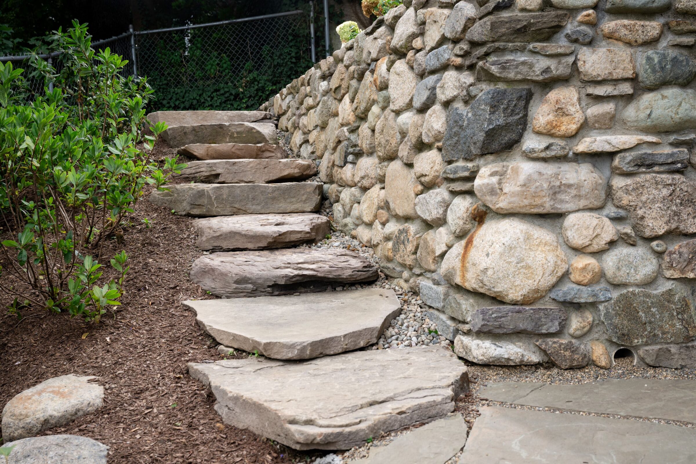 Stone steps lead up alongside a rustic, rock wall. Green shrubs grow nearby, next to a fence in a garden setting.