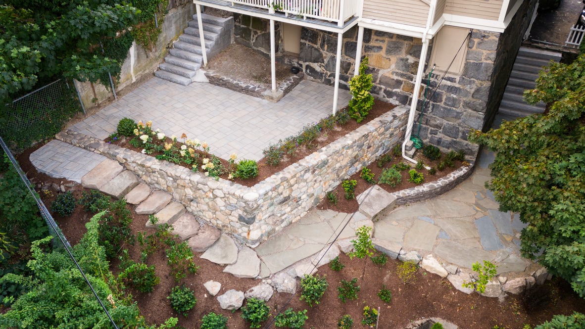 A terraced garden with stone walls, steps, and a patio, adjacent to a building. Lush greenery surrounds the well-maintained space.