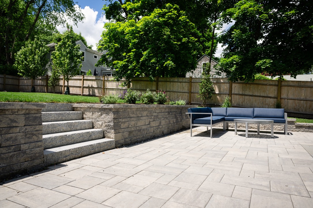 A backyard patio with stone steps, outdoor seating, and a wooden fence, surrounded by lush green trees and shrubs on a sunny day.