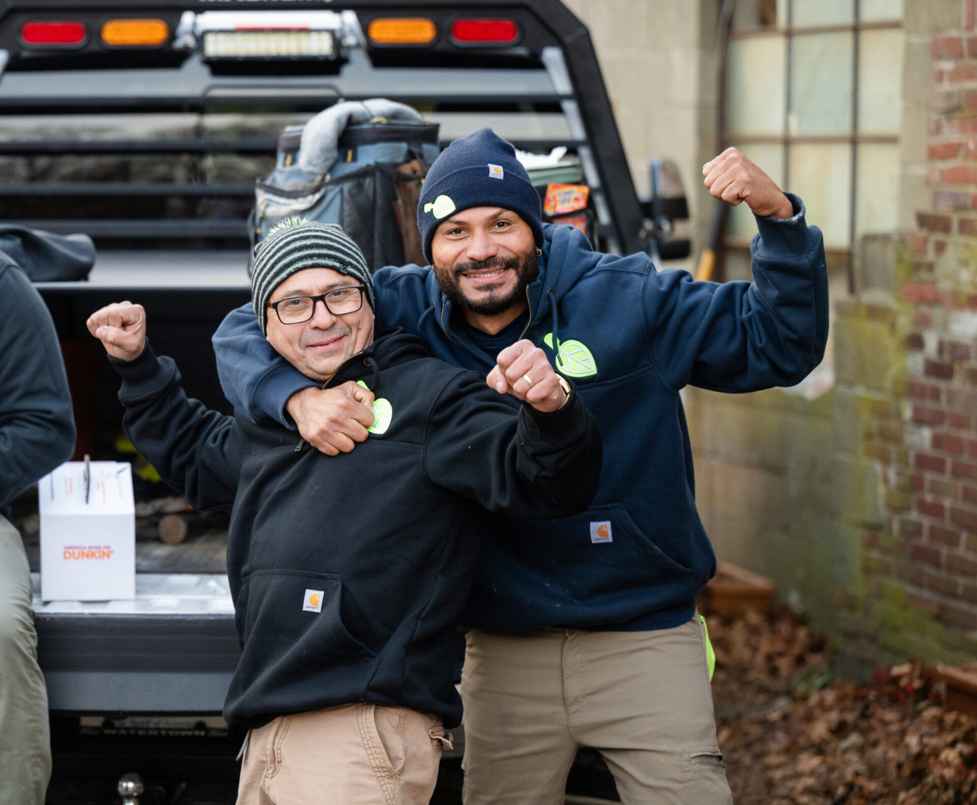 Two smiling people in casual clothing pose playfully near a vehicle. One person wears a beanie. A Dunkin’ box sits on the vehicle.