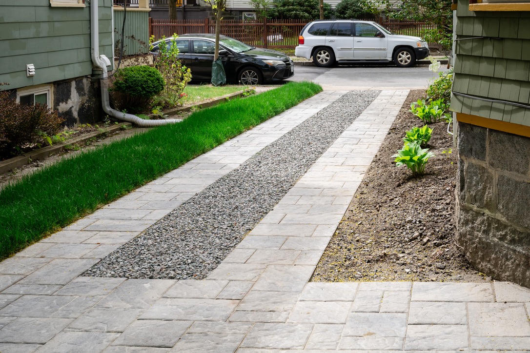 A neatly paved driveway with gravel center, surrounded by green grass, leads to the street. Two cars are parked nearby.