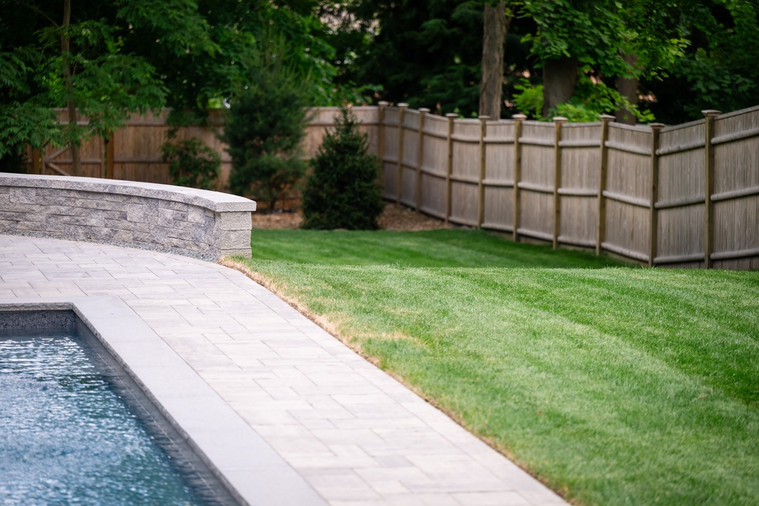 A tranquil backyard with a stone patio, swimming pool, green lawn, wooden fence, and lush trees creating a peaceful outdoor space.