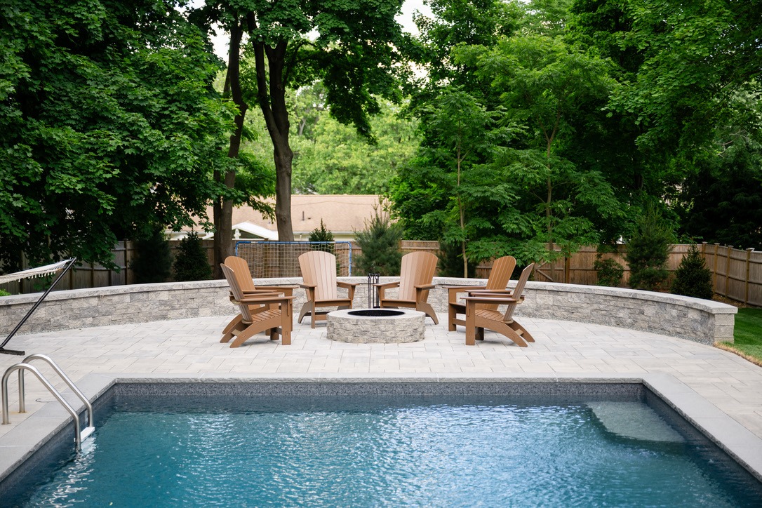 A serene backyard with a pool, surrounded by trees. Wooden chairs encircle a fire pit on a stone patio.