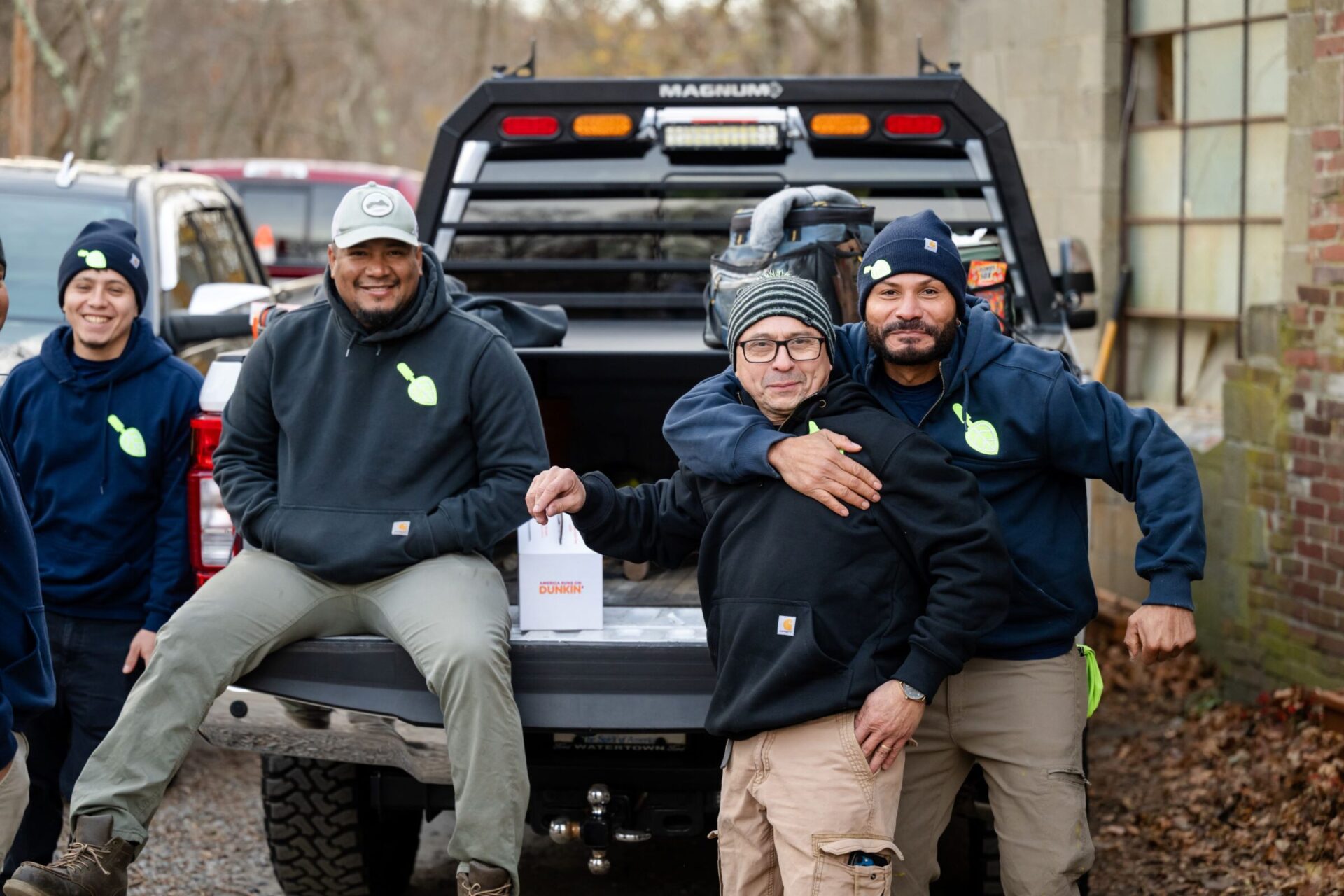 Three people in hoodies near a truck, smiling. One person holds a Dunkin' bag. Outdoors with parked vehicles in the background.