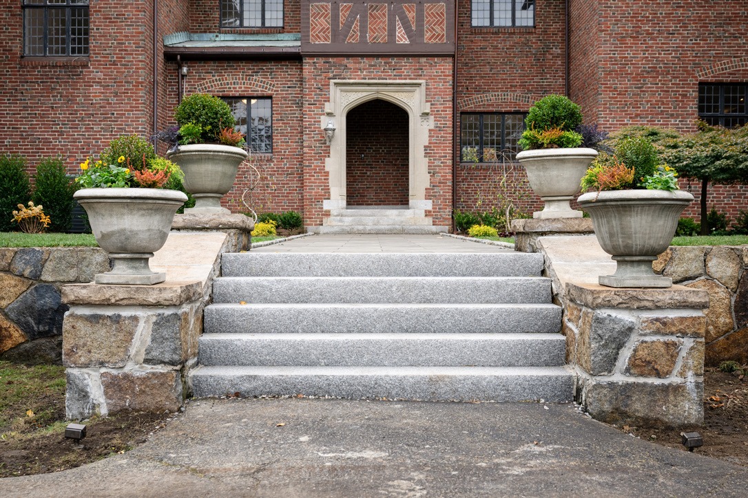 A brick building entrance with stone steps, large planters filled with greenery, and ornate archway, framed by manicured shrubs.