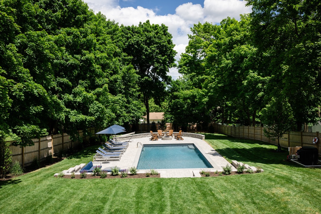 A backyard with a rectangular pool, surrounded by loungers, green grass, tall trees, and a wooden fence under a partly cloudy sky.