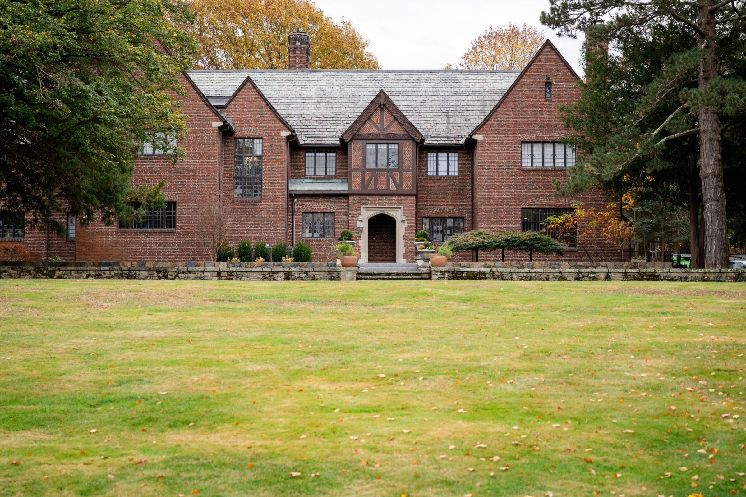 A large brick Tudor-style building with a spacious lawn, surrounded by trees and autumn foliage under an overcast sky.