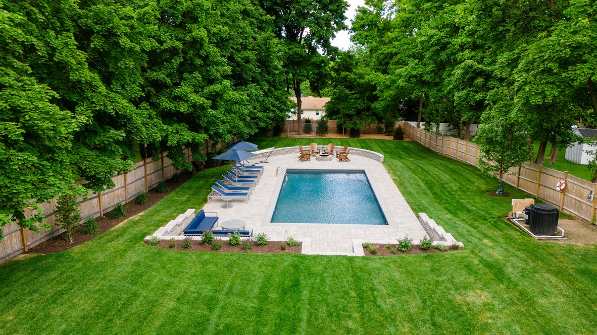 Aerial view of a backyard with a pool, surrounded by trees and a fence, featuring lounge chairs and a small seating area.