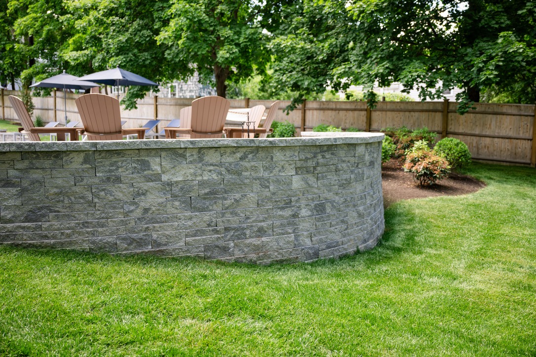 A serene backyard features wooden chairs, umbrellas, stone wall, lush green grass, and trees under clear skies, providing a tranquil outdoor setting.