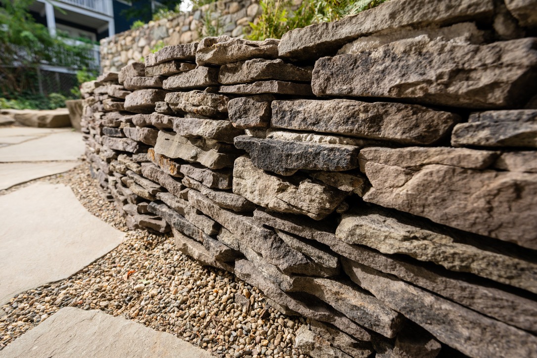 Close-up of a rustic stone wall with layered rocks, surrounded by gravel and plants, providing a natural, textured aesthetic.