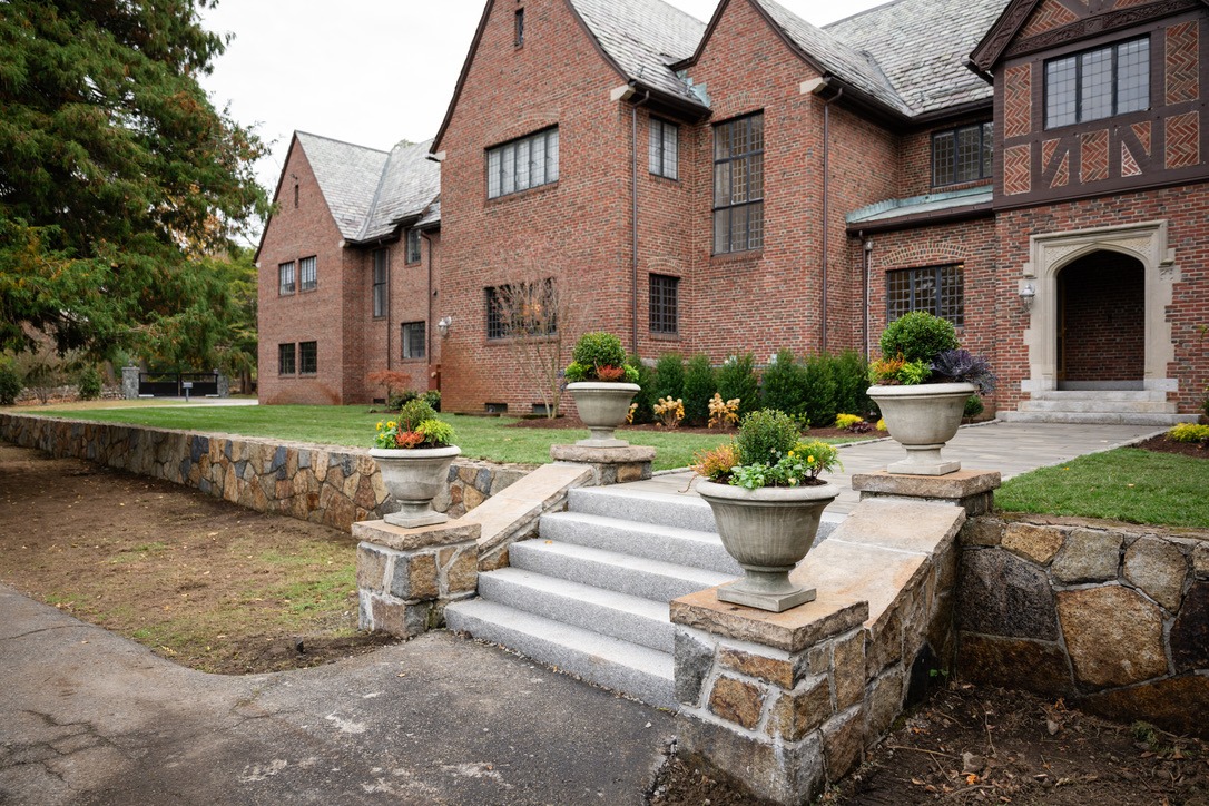 A historic brick building with stone steps and planters, surrounded by greenery. The architecture features Tudor-style design elements.