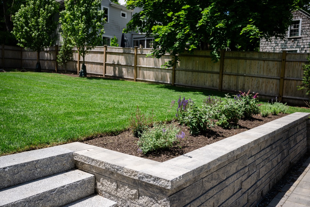 A well-maintained backyard features a lush lawn, raised stone flower bed with purple blooms, and a wooden fence surrounding the area.