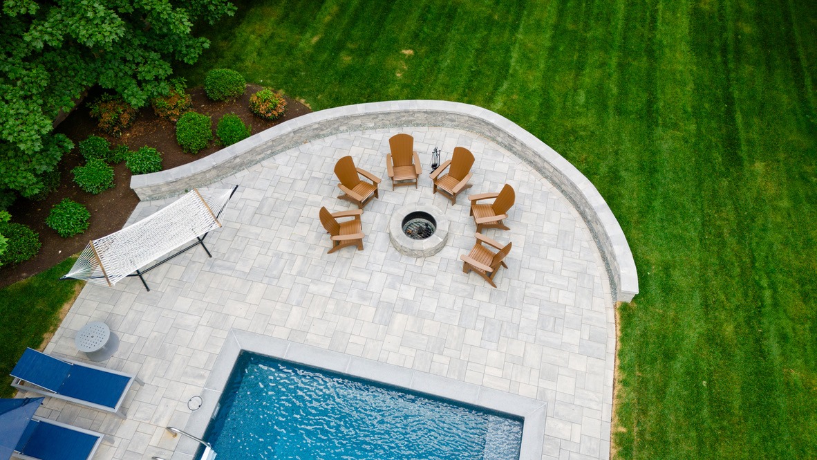 Aerial view of a backyard featuring a circular fire pit with six chairs, a hammock, pool, and neatly landscaped green lawn.