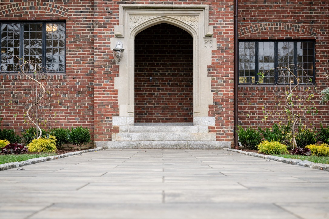 A paved walkway leads to a brick building entrance with arched stone details, surrounded by trimmed bushes and leafy plants.