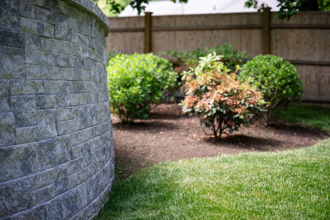 Stone wall beside a well-maintained garden with bushes and a wooden fence in the background on a sunny day.