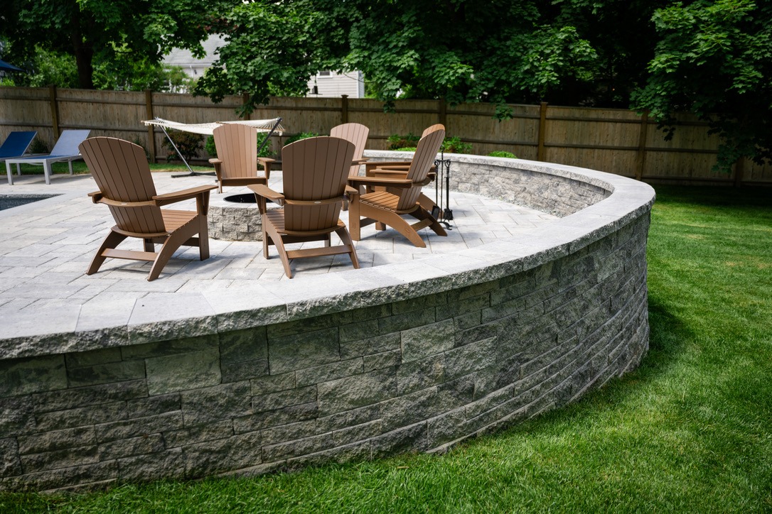 A curved stone patio with wooden chairs surrounds a fire pit, set in a grassy backyard with trees and a hammock.