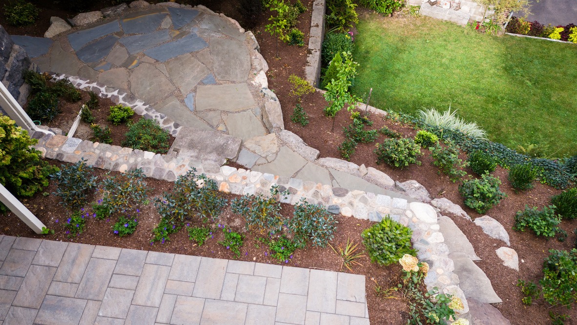 A stone pathway winds through a landscaped garden with lush plants, a grassy area, and decorative flowers, viewed from above.