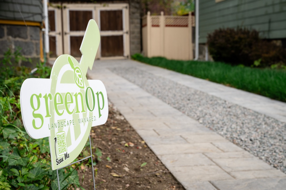 A landscaped garden path leads to wooden doors, featuring a "greenOp" sign for landscape management beside the walkway.