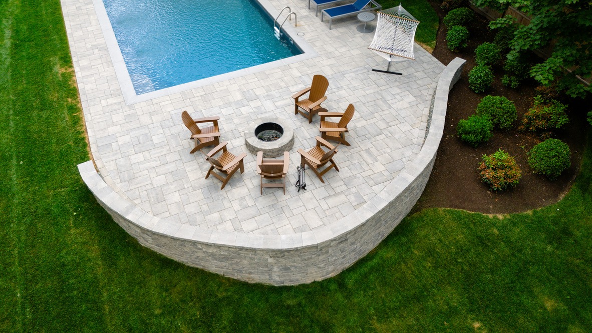 Aerial view of a backyard with a swimming pool, fire pit surrounded by chairs, hammock, and landscaped garden.